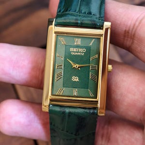 May include: A rectangular Seiko Quartz watch with a gold-tone case and a green dial. The watch features Roman numeral hour markers and gold hands. The watch has a green leather strap.