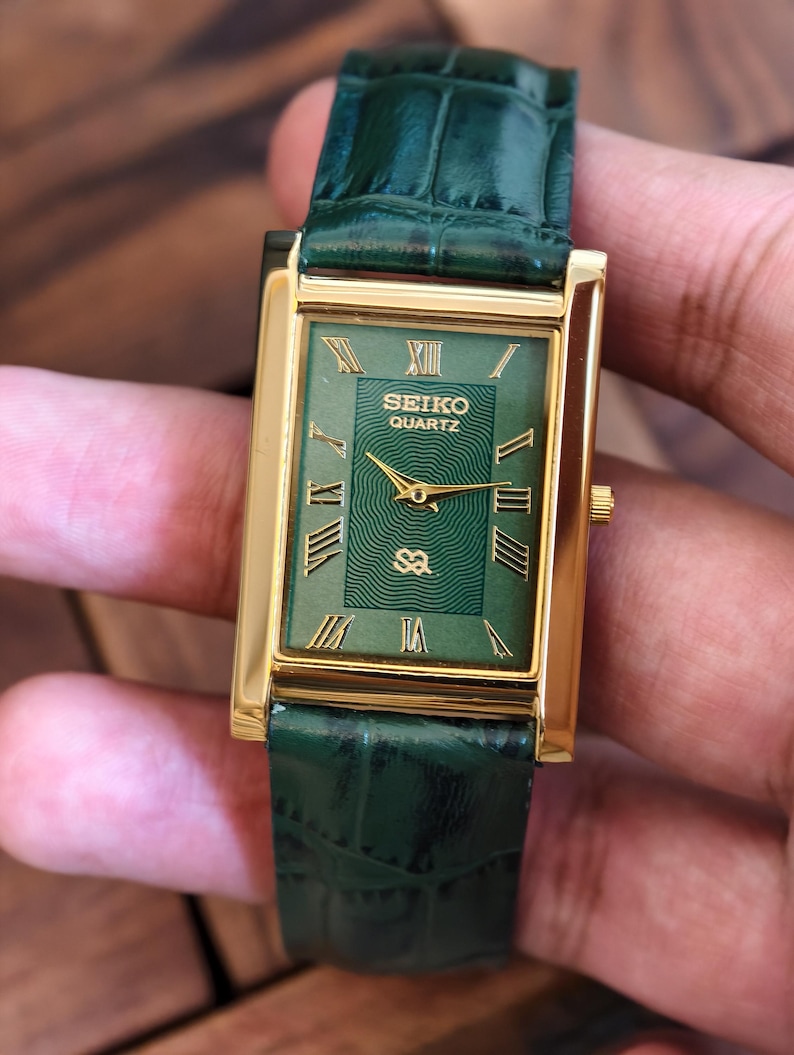 May include: A rectangular Seiko Quartz watch with a gold-tone case and a green dial. The watch features Roman numeral hour markers and gold hands. The watch has a green leather strap.