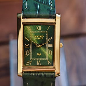 May include: A rectangular gold-tone Seiko Quartz watch with a green face and matching green leather band. The watch face features Roman numerals and the Seiko logo. The band has a crocodile skin texture.