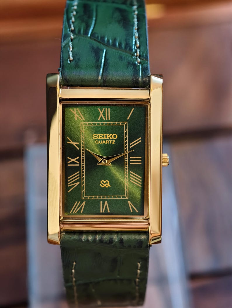 May include: A rectangular gold-tone Seiko Quartz watch with a green face and matching green leather band. The watch face features Roman numerals and the Seiko logo. The band has a crocodile skin texture.
