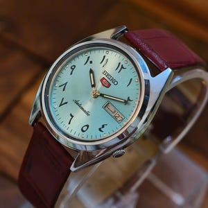 Pode incluir: Um relógio de pulso automático Seiko com mostrador azul claro e algarismos arábicos. O relógio tem caixa prateada e pulseira de couro bordô. A data é exibida às 3 horas. O relógio é exibido em um suporte transparente.