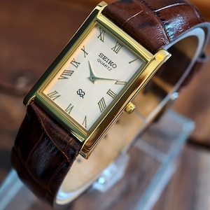 May include: A gold-toned Seiko Quartz watch with a rectangular face and brown leather band. The watch face is cream-colored with black Roman numerals and hands. The band has a textured, crocodile-like pattern.