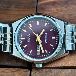 May include: A Seiko automatic wristwatch with a burgundy dial and silver-tone case and band. The watch face displays the day and date, with gold-tone hands and markers. The watch is marked "Japan Made" and "21 Jewels".
