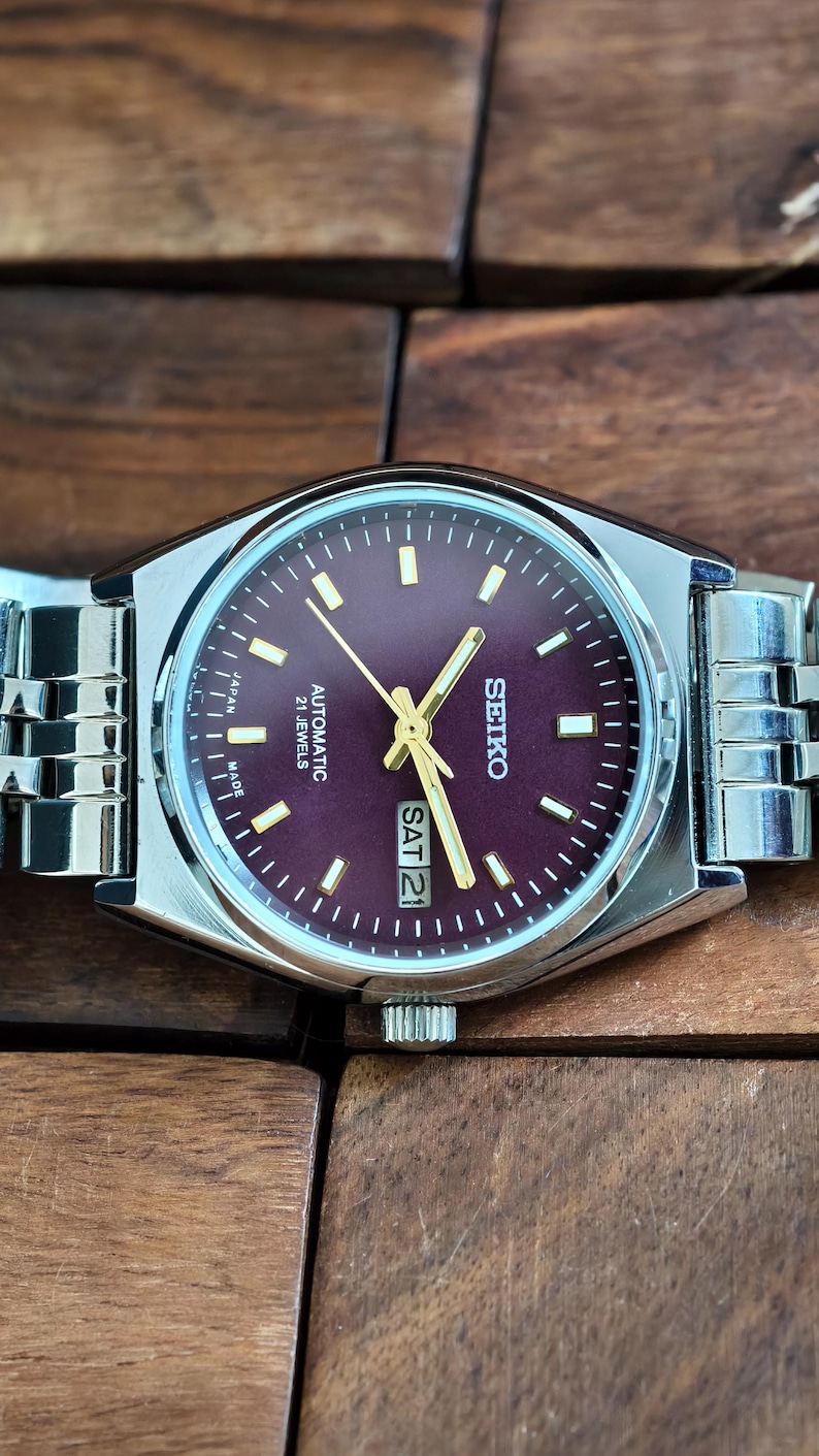 May include: A Seiko automatic wristwatch with a burgundy dial and silver-tone case and band. The watch face displays the day and date, with gold-tone hands and markers. The watch is marked "Japan Made" and "21 Jewels".