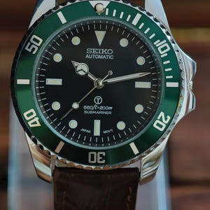 K&ouml;nnte beinhalten: Nahaufnahme einer Seiko Automatic Submariner Uhr mit schwarzem Zifferblatt und gr&uuml;ner L&uuml;nette. Die Uhr hat ein Edelstahlgeh&auml;use und ein braunes Lederarmband. Das Zifferblatt zeigt die Zeit mit wei&szlig;en Markierungen und Zeigern an. Wasserdicht bis 200m.