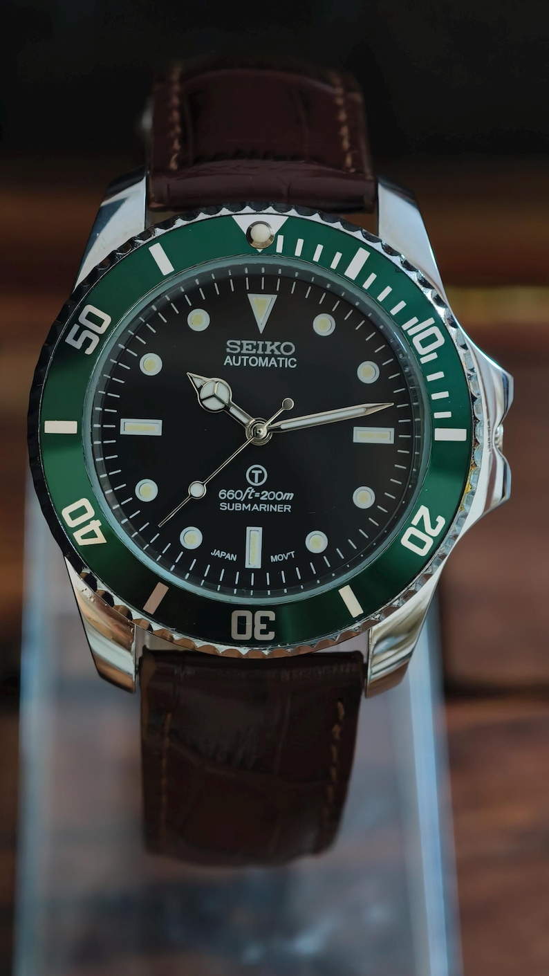 K&ouml;nnte beinhalten: Nahaufnahme einer Seiko Automatic Submariner Uhr mit schwarzem Zifferblatt und gr&uuml;ner L&uuml;nette. Die Uhr hat ein Edelstahlgeh&auml;use und ein braunes Lederarmband. Das Zifferblatt zeigt die Zeit mit wei&szlig;en Markierungen und Zeigern an. Wasserdicht bis 200m.
