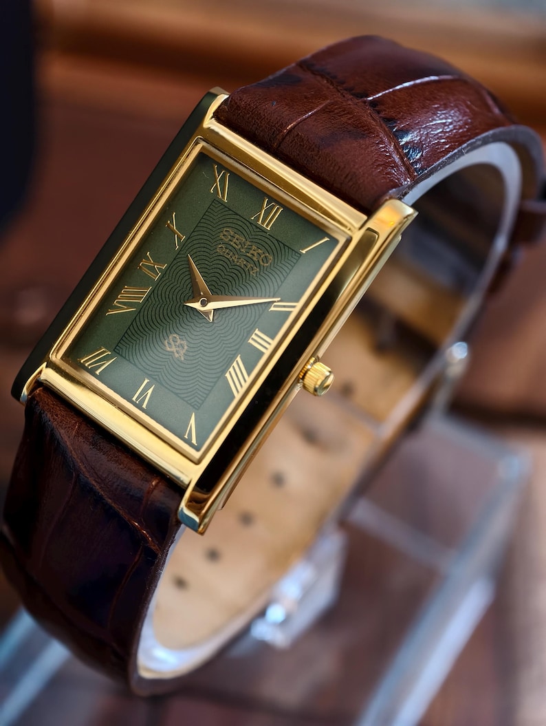 May include: A rectangular gold-plated wristwatch with a green face and Roman numeral hour markers. The watch has gold hands and a brown leather strap. The watch face has a textured pattern. The brand name is visible.
