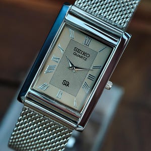 May include: A rectangular Seiko Quartz watch with a silver mesh strap. The watch face is a light beige colour with Roman numerals and the Seiko Quartz logo. The watch has a silver case and a small crown. The watch is displayed on a clear stand.
