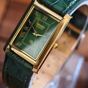 May include: A rectangular Seiko Quartz watch with a gold-tone case and a green dial. The watch features Roman numeral hour markers and gold hands. The watch has a green leather band with a crocodile pattern.