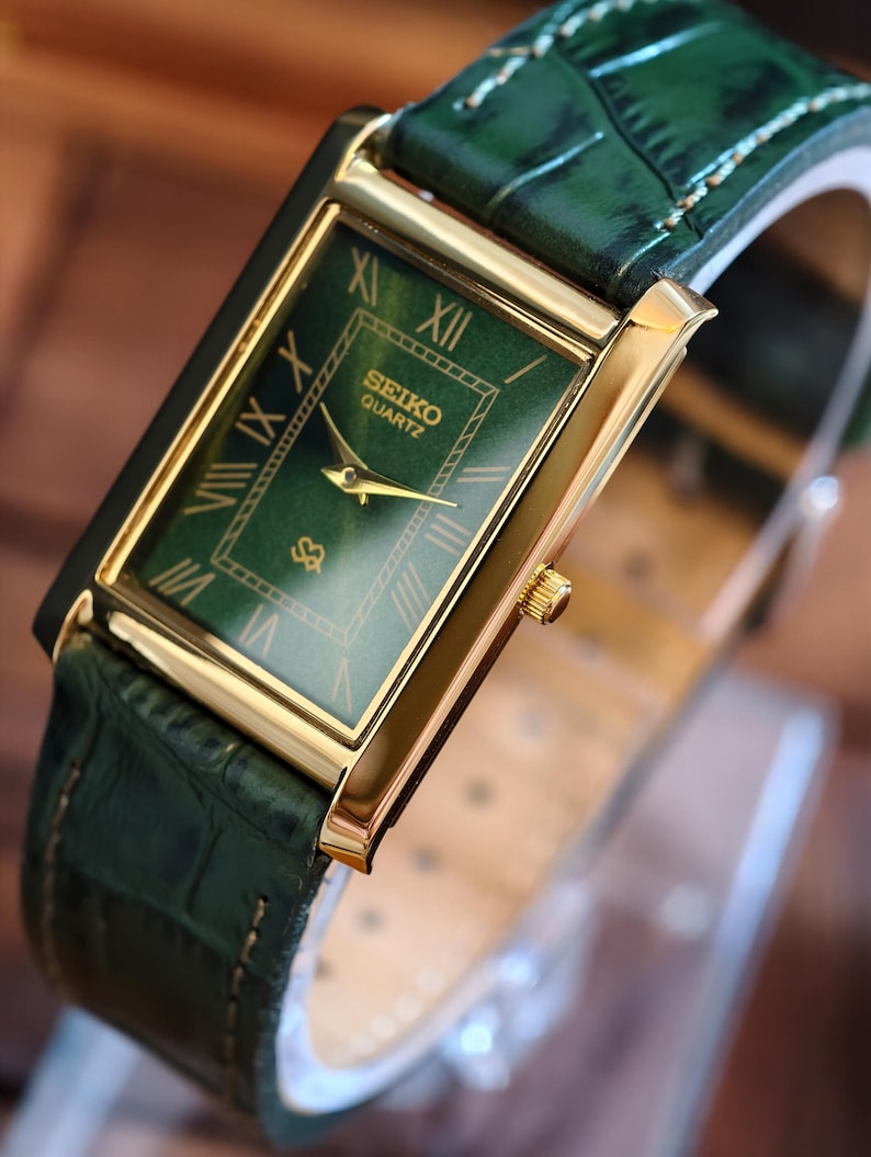 May include: A rectangular Seiko Quartz watch with a gold-tone case and a green dial. The watch features Roman numeral hour markers and gold hands. The watch has a green leather band with a crocodile pattern.