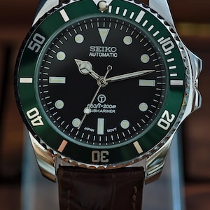 K&ouml;nnte beinhalten: Eine Seiko Automatic Submariner Uhr mit schwarzem Zifferblatt und gr&uuml;ner L&uuml;nette. Die Uhr hat ein Edelstahlgeh&auml;use und ein braunes Lederarmband. Das Zifferblatt hat Leuchtmarkierungen und Zeiger. Die Uhr ist bis 200 Meter wasserdicht.