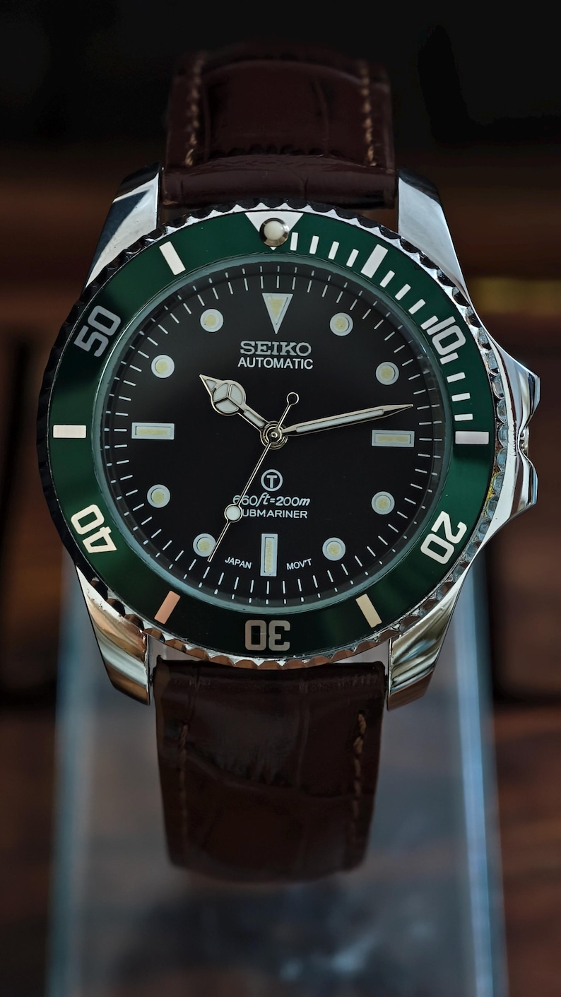 K&ouml;nnte beinhalten: Eine Seiko Automatic Submariner Uhr mit schwarzem Zifferblatt und gr&uuml;ner L&uuml;nette. Die Uhr hat ein Edelstahlgeh&auml;use und ein braunes Lederarmband. Das Zifferblatt hat Leuchtmarkierungen und Zeiger. Die Uhr ist bis 200 Meter wasserdicht.