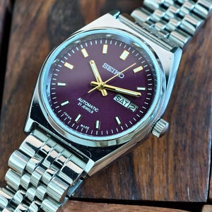 May include: A silver-toned Seiko automatic wristwatch with a deep purple dial. The watch features gold-colored hands and markers, a day-date display, and a stainless steel band. The watch is displayed on a wooden surface.