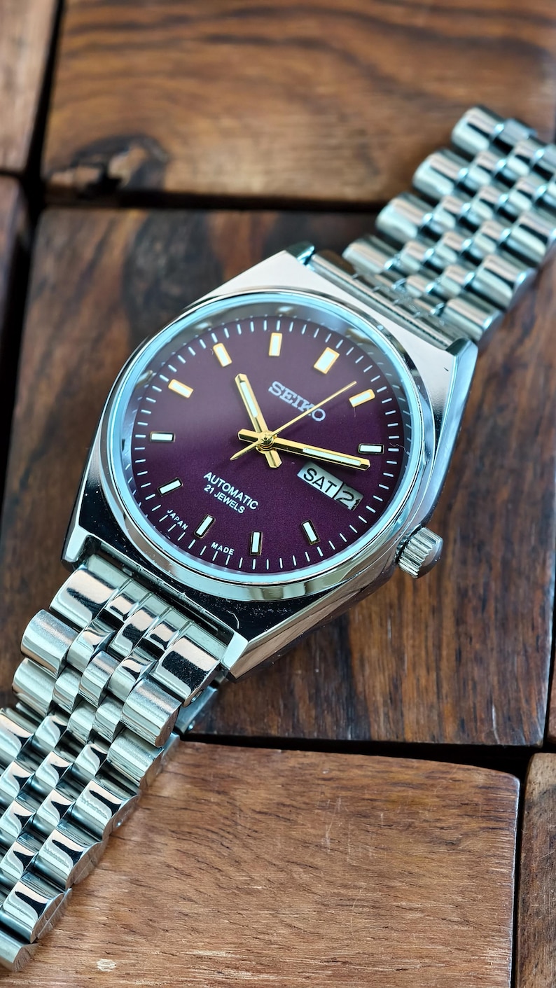 May include: A silver-toned Seiko automatic wristwatch with a deep purple dial. The watch features gold-colored hands and markers, a day-date display, and a stainless steel band. The watch is displayed on a wooden surface.