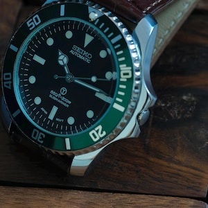 K&ouml;nnte beinhalten: Nahaufnahme einer Seiko Automatic Submariner Uhr mit schwarzem Zifferblatt und gr&uuml;ner L&uuml;nette. Die Uhr hat ein Edelstahlgeh&auml;use und ein braunes Lederarmband. Das Zifferblatt zeigt den Text "660ft/200m Submariner".
