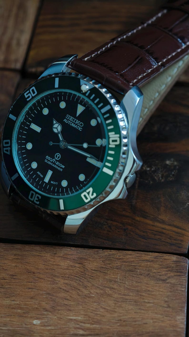 K&ouml;nnte beinhalten: Nahaufnahme einer Seiko Automatic Submariner Uhr mit schwarzem Zifferblatt und gr&uuml;ner L&uuml;nette. Die Uhr hat ein Edelstahlgeh&auml;use und ein braunes Lederarmband. Das Zifferblatt zeigt den Text "660ft/200m Submariner".