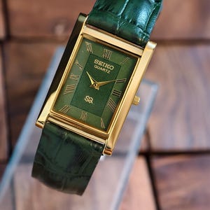 May include: A rectangular Seiko Quartz watch with a gold-tone case and a green dial. The watch features Roman numeral hour markers and gold hands. The green leather strap complements the watch face. The watch is displayed on a clear surface.