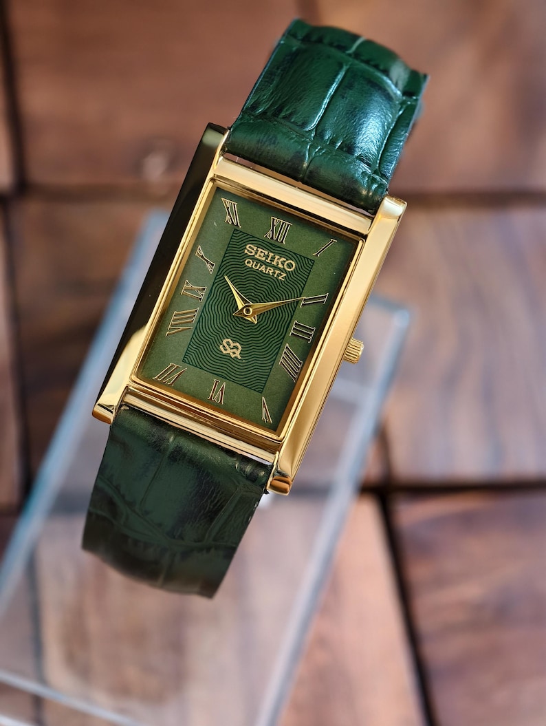 May include: A rectangular Seiko Quartz watch with a gold-tone case and a green dial. The watch features Roman numeral hour markers and gold hands. The green leather strap complements the watch face. The watch is displayed on a clear surface.