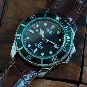 K&ouml;nnte beinhalten: Eine Seiko Automatic Submariner Uhr mit schwarzem Zifferblatt und gr&uuml;ner L&uuml;nette. Die Uhr hat ein Edelstahlgeh&auml;use und ein braunes Lederarmband. Das Zifferblatt zeigt die Zeit mit wei&szlig;en Markierungen und Zeigern an. Die Uhr ist wasserdicht bis 200m.