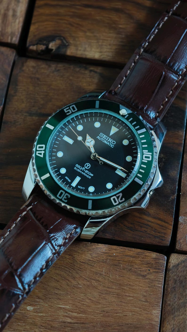 K&ouml;nnte beinhalten: Eine Seiko Automatic Submariner Uhr mit schwarzem Zifferblatt und gr&uuml;ner L&uuml;nette. Die Uhr hat ein Edelstahlgeh&auml;use und ein braunes Lederarmband. Das Zifferblatt zeigt die Zeit mit wei&szlig;en Markierungen und Zeigern an. Die Uhr ist wasserdicht bis 200m.