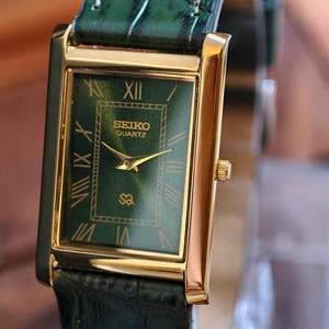 May include: A gold-toned Seiko Quartz watch with a rectangular green face and Roman numeral hour markers. The watch features a green leather band. The watch face displays the Seiko Quartz logo and gold hands.