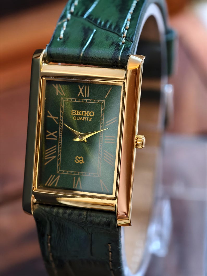 May include: A gold-toned Seiko Quartz watch with a rectangular green face and Roman numeral hour markers. The watch features a green leather band. The watch face displays the Seiko Quartz logo and gold hands.
