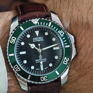 K&ouml;nnte beinhalten: Seiko Automatic Submariner Uhr mit schwarzem Zifferblatt und gr&uuml;ner L&uuml;nette. Die Uhr hat ein Edelstahlgeh&auml;use und ein braunes Lederarmband. Das Zifferblatt zeigt die Zeit mit wei&szlig;en Markierungen und Zeigern an.