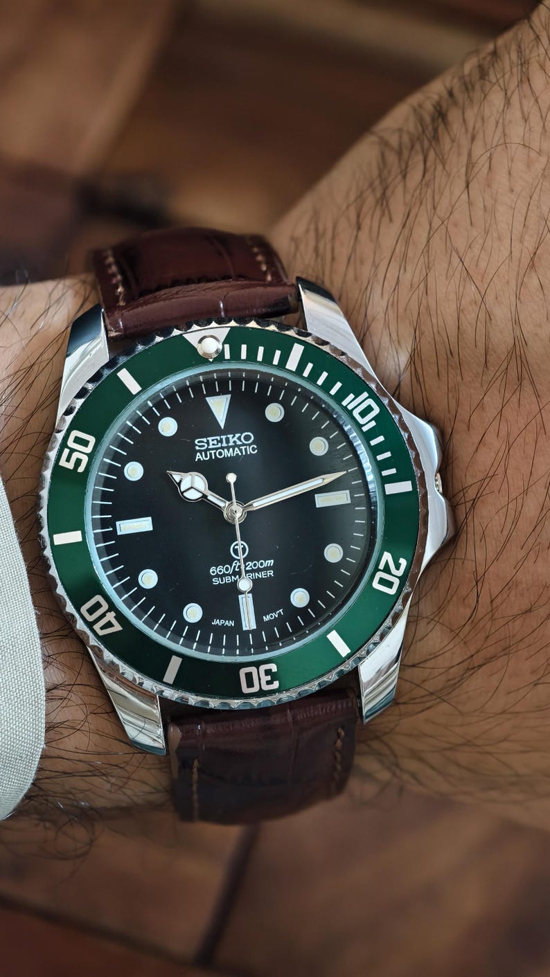 K&ouml;nnte beinhalten: Seiko Automatic Submariner Uhr mit schwarzem Zifferblatt und gr&uuml;ner L&uuml;nette. Die Uhr hat ein Edelstahlgeh&auml;use und ein braunes Lederarmband. Das Zifferblatt zeigt die Zeit mit wei&szlig;en Markierungen und Zeigern an.