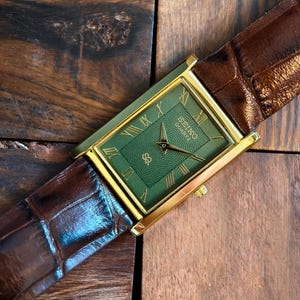 May include: A gold rectangular watch with a green face and gold Roman numerals. The watch has a brown leather strap. The watch face has the text "SEIKO QUARTZ".