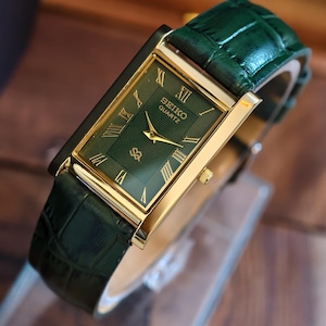 May include: A gold-plated Seiko Quartz watch with a green face and Roman numerals. The watch has a green leather strap. The watch face has a textured pattern and the Seiko logo. The watch's dimensions are approximately 3.5 cm x 2.5 cm.