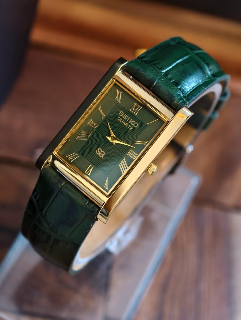 May include: A gold-plated Seiko Quartz watch with a green face and Roman numerals. The watch has a green leather strap. The watch face has a textured pattern and the Seiko logo. The watch's dimensions are approximately 3.5 cm x 2.5 cm.