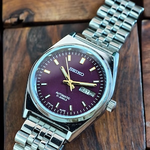 May include: A silver-toned Seiko automatic wristwatch with a burgundy dial and a stainless steel band. The watch face displays the day and date, with gold-colored hands and markers. The watch is marked "Made in Japan."