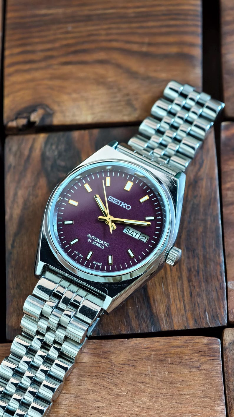 May include: A silver-toned Seiko automatic wristwatch with a burgundy dial and a stainless steel band. The watch face displays the day and date, with gold-colored hands and markers. The watch is marked "Made in Japan."