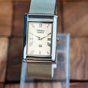 Puede incluir: Un reloj Seiko Quartz rectangular con una correa de malla plateada. La esfera del reloj es de color crema con números romanos y manecillas negras. El reloj tiene una caja plateada y una pequeña corona en el lateral. El reloj se exhibe sobre un soporte transparente.