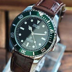 K&ouml;nnte beinhalten: Eine Seiko Automatic Uhr mit schwarzem Zifferblatt und gr&uuml;ner L&uuml;nette. Die Uhr hat ein braunes Lederarmband und ein silbernes Geh&auml;use. Das Zifferblatt zeigt den Text "SEIKO AUTOMATIC" und "660ft=200m SUBMARINER".