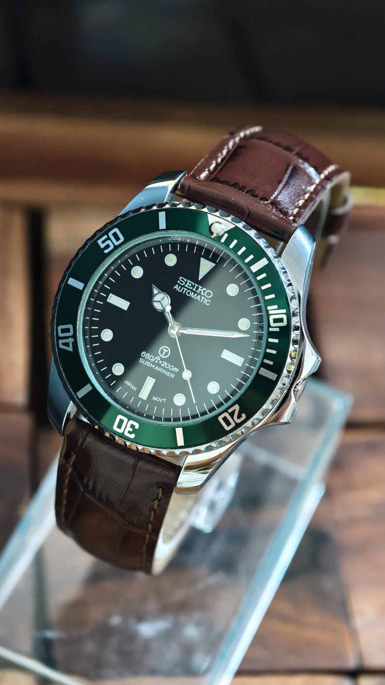 K&ouml;nnte beinhalten: Eine Seiko Automatic Uhr mit schwarzem Zifferblatt und gr&uuml;ner L&uuml;nette. Die Uhr hat ein braunes Lederarmband und ein silbernes Geh&auml;use. Das Zifferblatt zeigt den Text "SEIKO AUTOMATIC" und "660ft=200m SUBMARINER".
