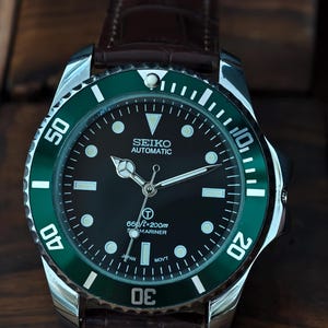 K&ouml;nnte beinhalten: Seiko Automatic Uhr mit schwarzem Zifferblatt und gr&uuml;ner L&uuml;nette. Die Uhr hat ein Edelstahlgeh&auml;use und ein braunes Lederarmband. Das Zifferblatt zeigt die Zeit mit wei&szlig;en Markierungen und Zeigern an. Wasserdicht bis 200 Meter.