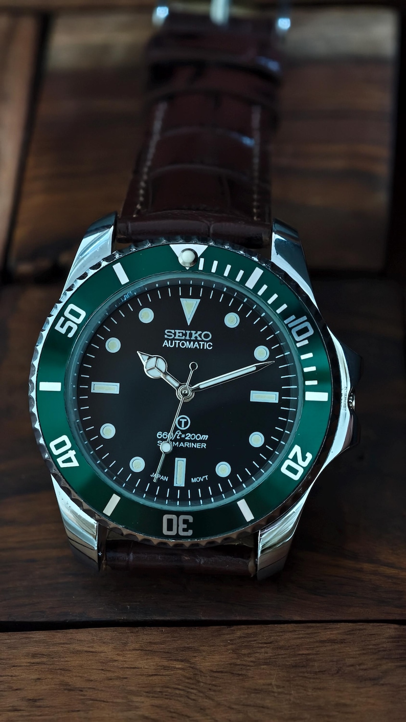 K&ouml;nnte beinhalten: Seiko Automatic Uhr mit schwarzem Zifferblatt und gr&uuml;ner L&uuml;nette. Die Uhr hat ein Edelstahlgeh&auml;use und ein braunes Lederarmband. Das Zifferblatt zeigt die Zeit mit wei&szlig;en Markierungen und Zeigern an. Wasserdicht bis 200 Meter.