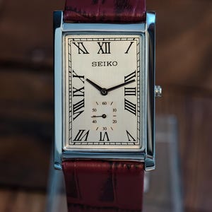 Op de afbeelding: Een rechthoekig Seiko-polshorloge met een zilveren kast en een crèmekleurige wijzerplaat. De wijzerplaat heeft zwarte Romeinse cijfers en wijzers, met de merknaam "SEIKO". De band is van bordeauxrood leer.