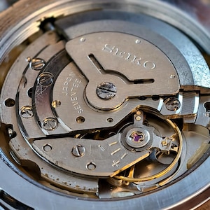 K&ouml;nnte beinhalten: Nahaufnahme eines Seiko-Uhrwerks, das komplizierte mechanische Komponenten zeigt. Die silberfarbenen Zahnr&auml;der, Schrauben und Platten sind sichtbar, mit dem Wort "SEIKO" prominent dargestellt. Das Bild hebt die Pr&auml;zision und Handwerkskunst des Innenlebens der Uhr hervor.
