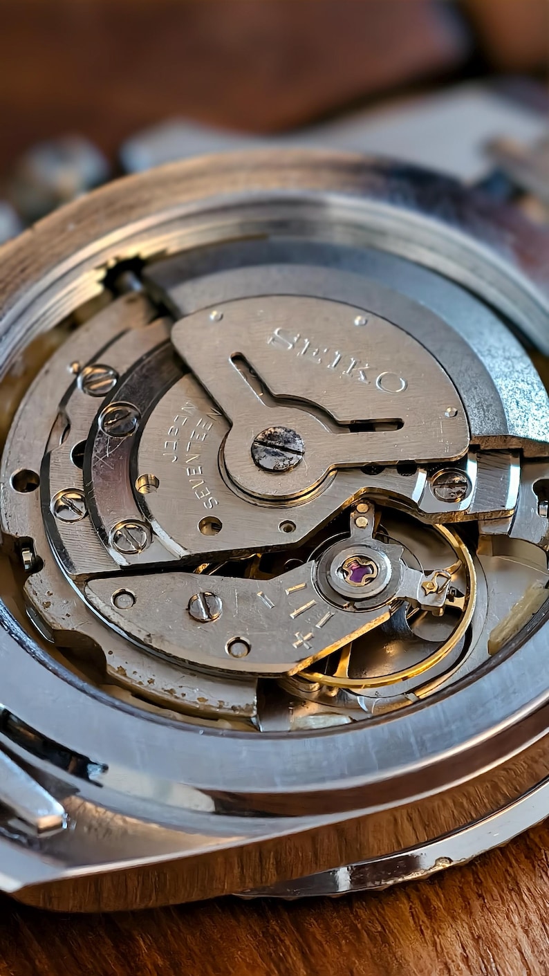 K&ouml;nnte beinhalten: Nahaufnahme eines Seiko-Uhrwerks, das komplizierte mechanische Komponenten zeigt. Die silberfarbenen Zahnr&auml;der, Schrauben und Platten sind sichtbar, mit dem Wort "SEIKO" prominent dargestellt. Das Bild hebt die Pr&auml;zision und Handwerkskunst des Innenlebens der Uhr hervor.