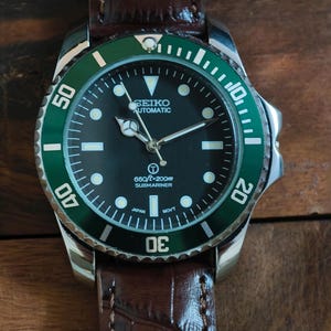 K&ouml;nnte beinhalten: Nahaufnahme einer Seiko Automatik-Uhr mit schwarzem Zifferblatt und gr&uuml;ner L&uuml;nette. Die Uhr hat ein braunes Lederarmband. Auf dem Zifferblatt steht "SEIKO AUTOMATIC" und "660ft/200m SUBMARINER".