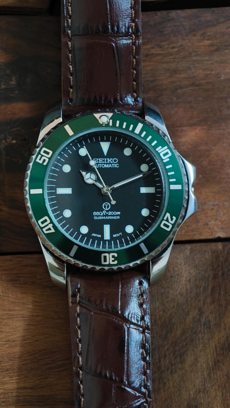 K&ouml;nnte beinhalten: Nahaufnahme einer Seiko Automatik-Uhr mit schwarzem Zifferblatt und gr&uuml;ner L&uuml;nette. Die Uhr hat ein braunes Lederarmband. Auf dem Zifferblatt steht "SEIKO AUTOMATIC" und "660ft/200m SUBMARINER".