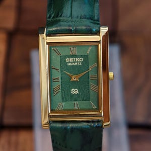 May include: A gold-cased Seiko Quartz watch with a green dial and a green leather strap. The watch face displays Roman numerals and gold hands. The rectangular watch has a classic design. The strap is made of green leather.