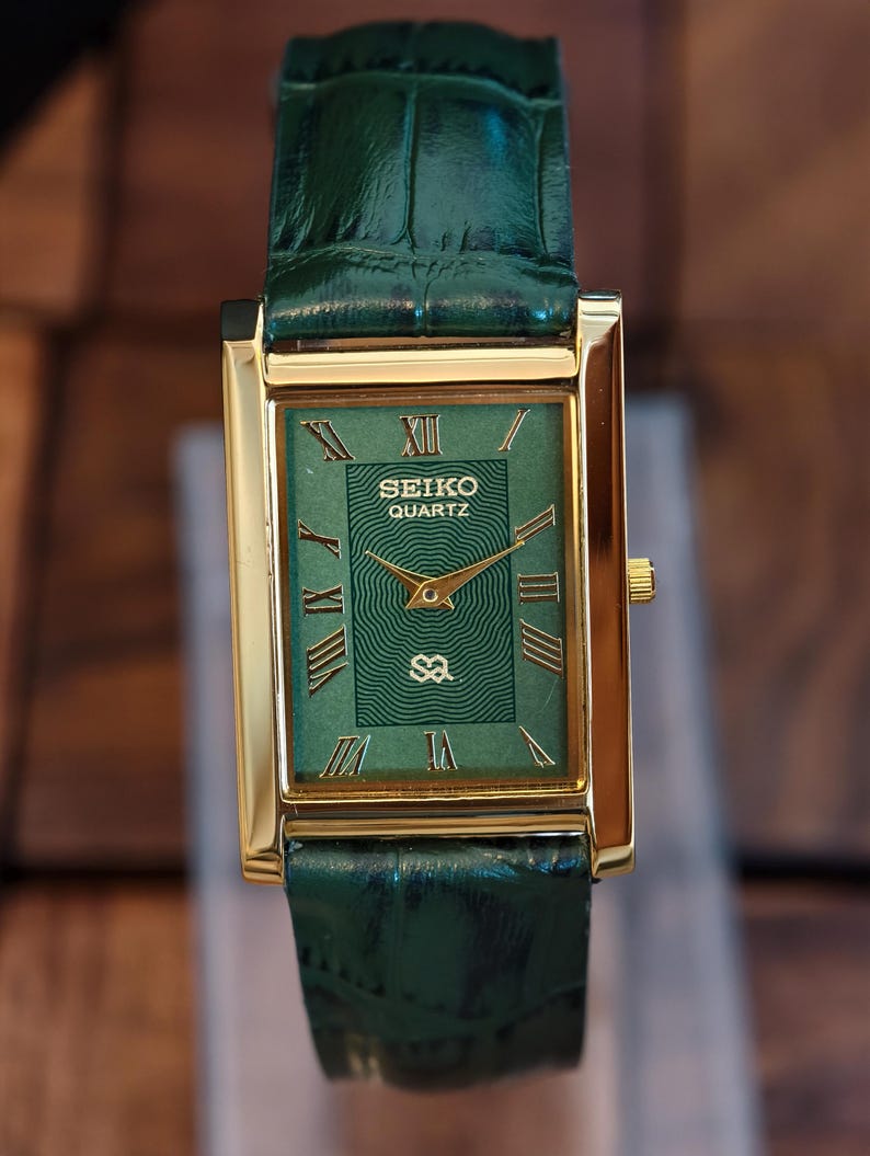 May include: A gold-cased Seiko Quartz watch with a green dial and a green leather strap. The watch face displays Roman numerals and gold hands. The rectangular watch has a classic design. The strap is made of green leather.