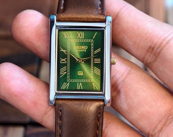 May include: A rectangular Seiko Quartz watch with a green face and Roman numeral hour markers. The watch has a silver-colored case and a brown leather band. The watch face has the Seiko Quartz logo and a gold-colored crown.
