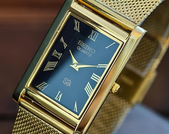 Può includere: Un orologio Seiko Quartz dorato con quadrante nero rettangolare e numeri romani dorati. L'orologio presenta un cinturino in maglia dorata e lancette dorate. Il quadrante mostra il logo Seiko Quartz. L'orologio è un design classico.