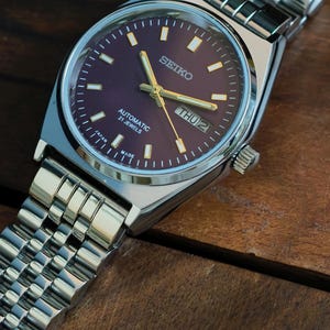 May include: A Seiko automatic wristwatch with a dark purple dial and silver-tone case and band. The watch face displays the day and date, with gold-tone hands and markers. The watch band is a silver, linked bracelet. The watch is labeled "Automatic 21 Jewels" and "Made in Japan."