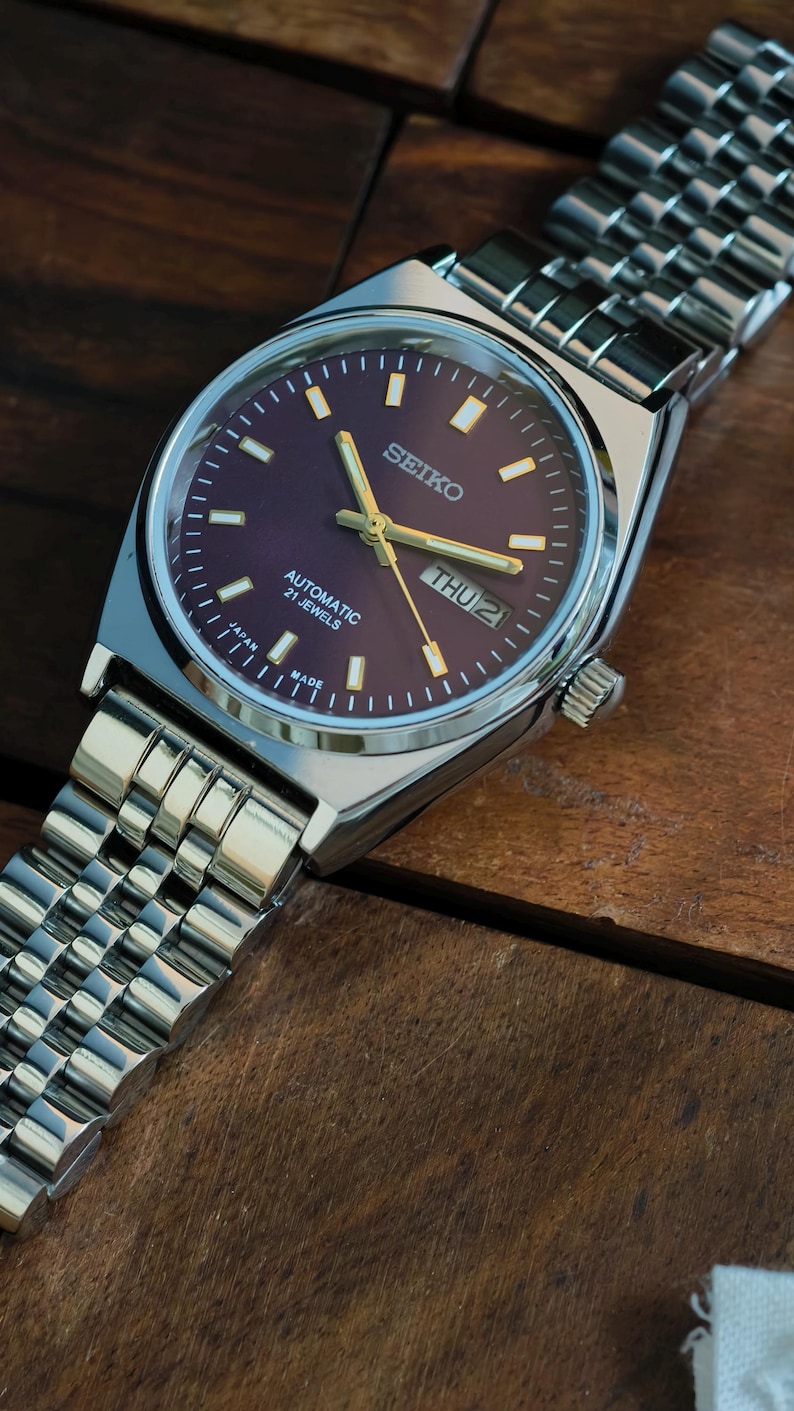 May include: A Seiko automatic wristwatch with a dark purple dial and silver-tone case and band. The watch face displays the day and date, with gold-tone hands and markers. The watch band is a silver, linked bracelet. The watch is labeled "Automatic 21 Jewels" and "Made in Japan."