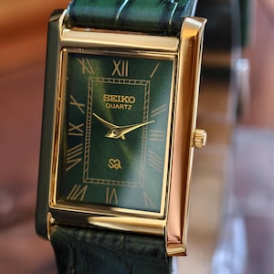 May include: A rectangular Seiko Quartz watch with a gold-tone case and a green dial. The watch features gold hands and Roman numeral hour markers. The watch has a green leather band.